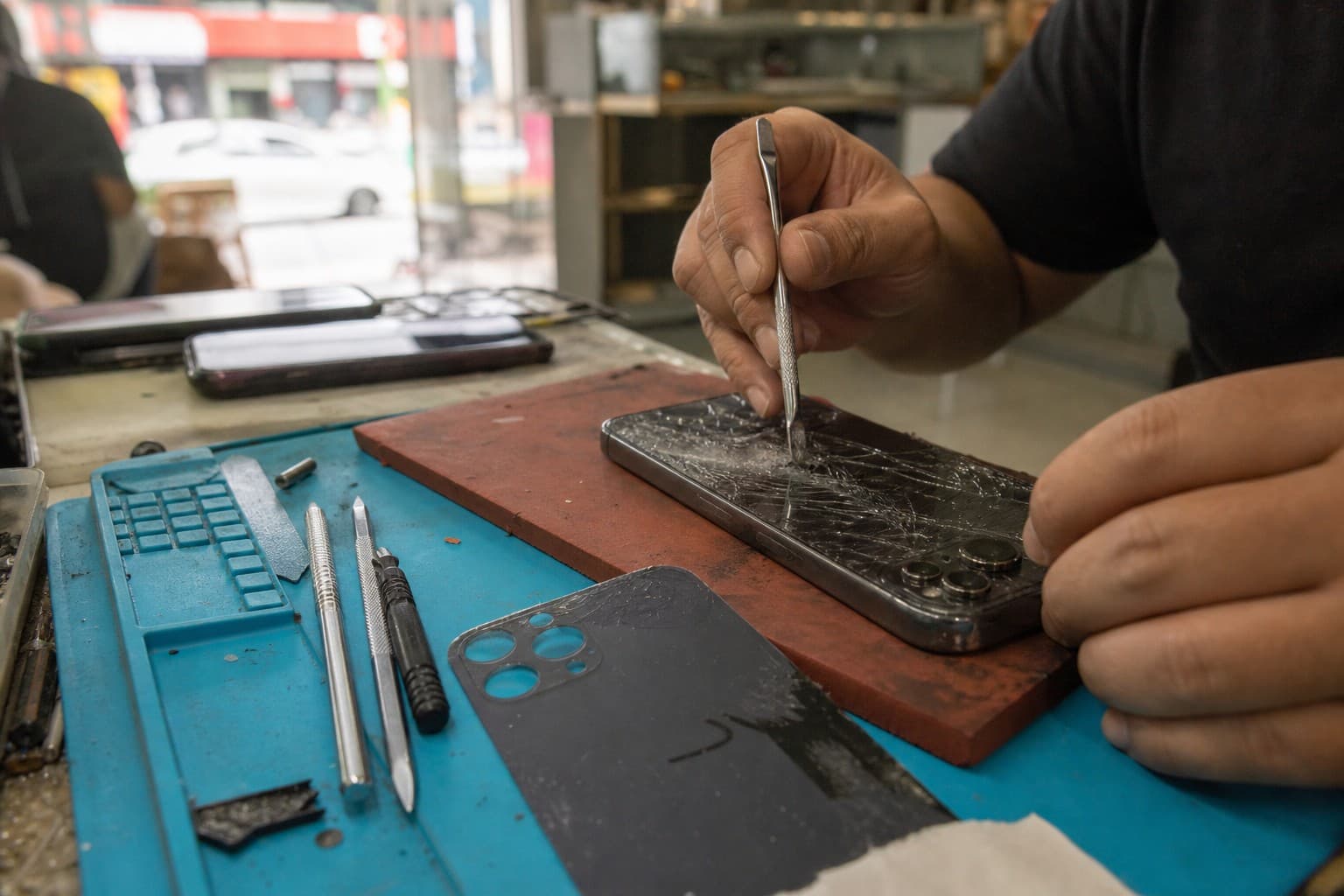 Technician working on the cracked rear glass of an iPhone on a repair-shop bench, a replacement back panel nearby