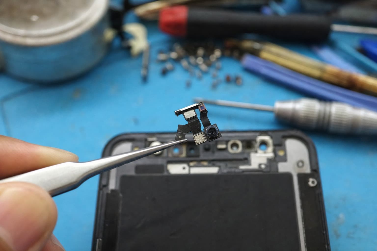 Close-up under a microscope of a technician using fine tweezers to handle a tiny front-camera and dot-projector flex cable from a smartphone on a repair-shop bench, on a blue anti-static mat