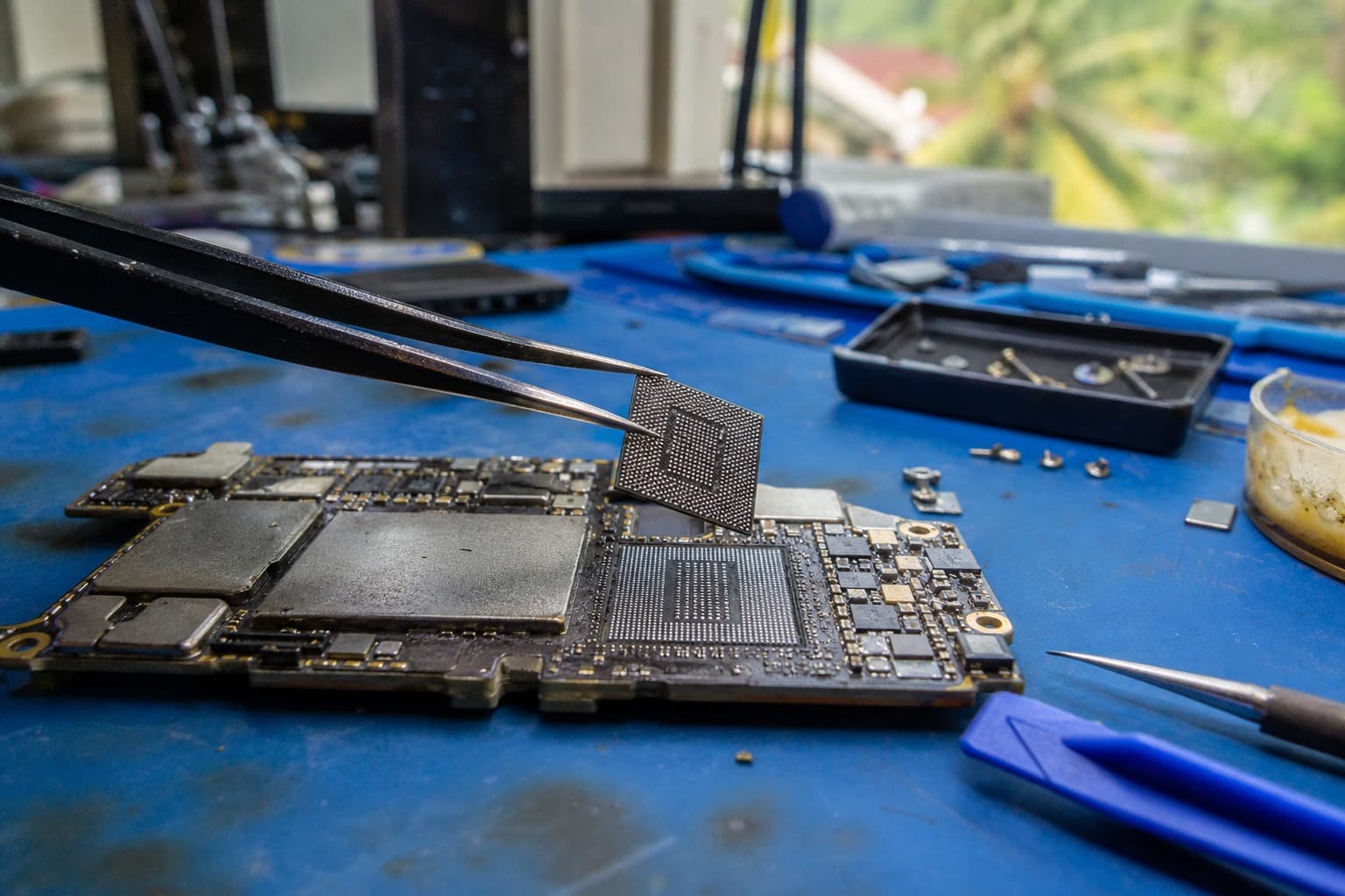 Phone logic board under rework with a storage chip lifted by tweezers, chip trays beside
