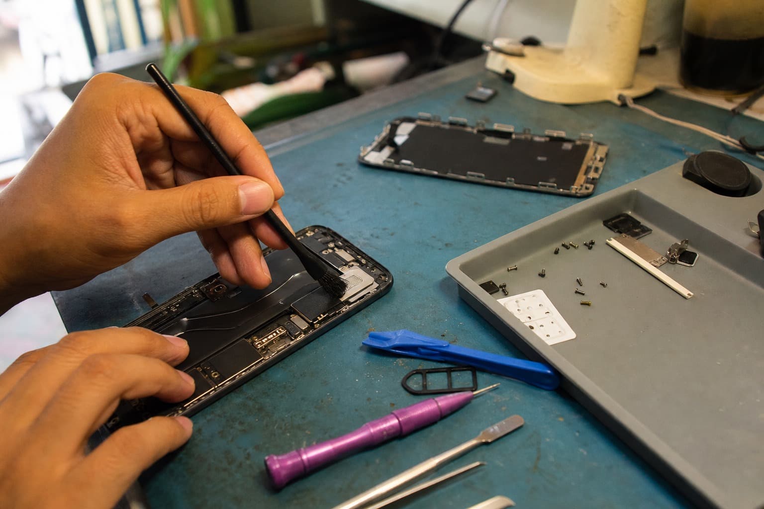 Hands cleaning the speaker mesh of a disassembled phone with an anti-static brush on a repair bench