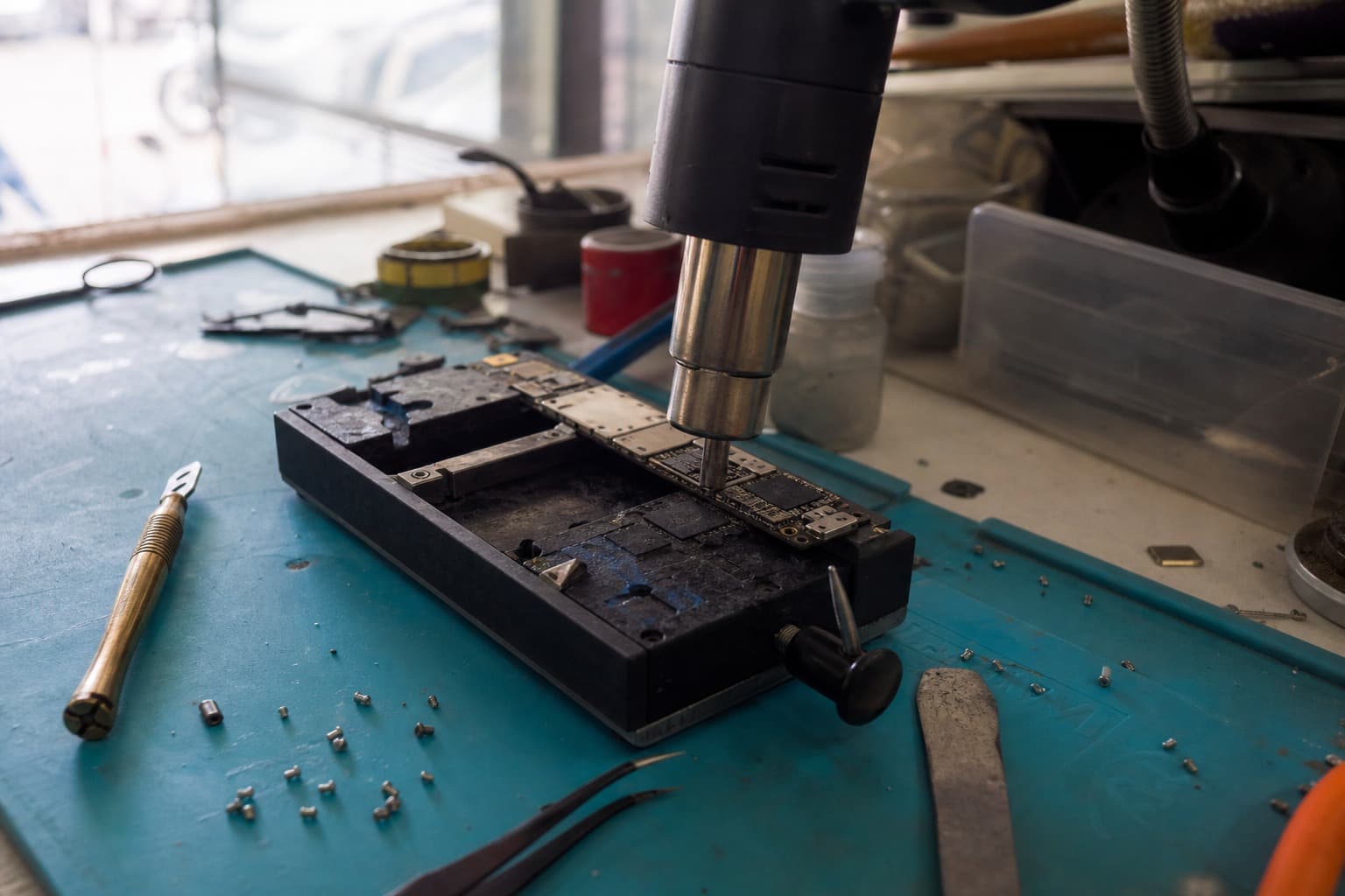 Phone logic board clamped on a repair bench with a hot-air rework station over the wireless chip area
