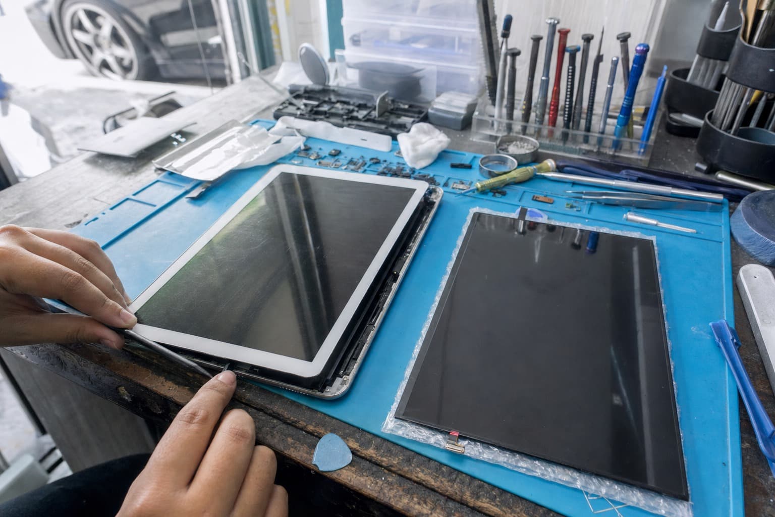 A large tablet on a repair bench with its front glass partially separated from the body by a thin pry tool, a replacement screen panel resting nearby on a blue anti-static mat, no logos