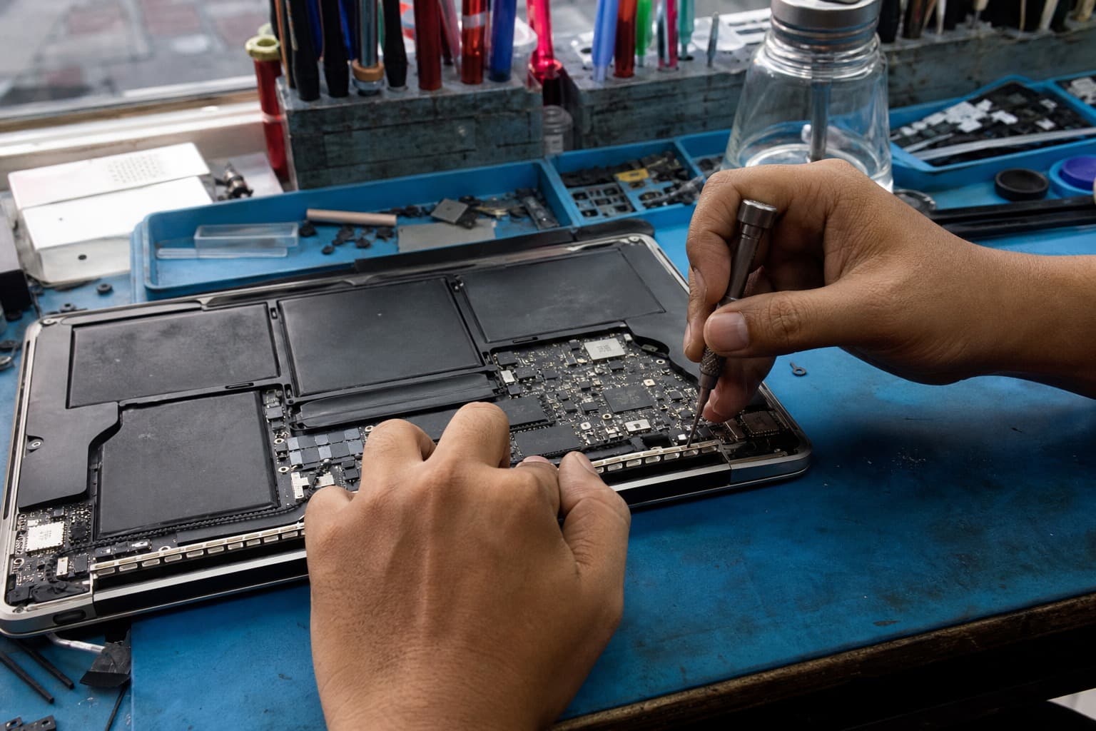 A thin aluminium laptop flipped upside-down on a repair bench with its bottom case removed, exposing the wide internal battery and logic board, a technician working with a precision screwdriver on a blue anti-static mat, no logos