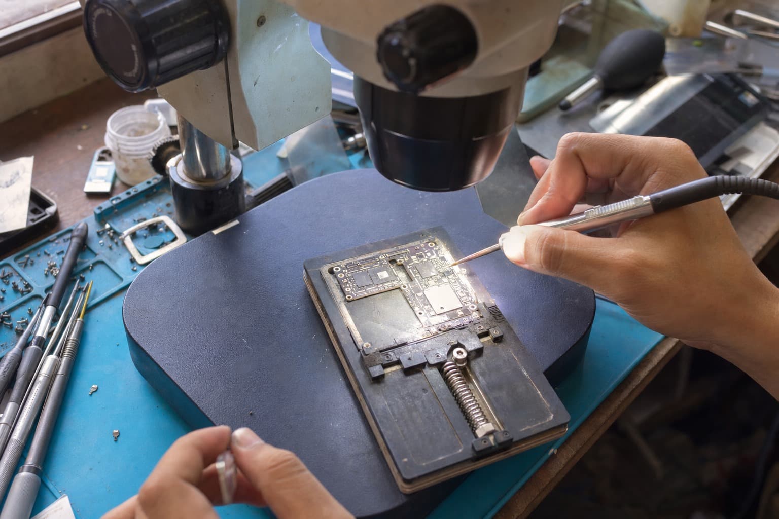 A OnePlus-style Android flagship logic board clamped under a microscope while a technician does micro-soldering rework on a tiny chip with a fine soldering tip, blue anti-static mat, no logos