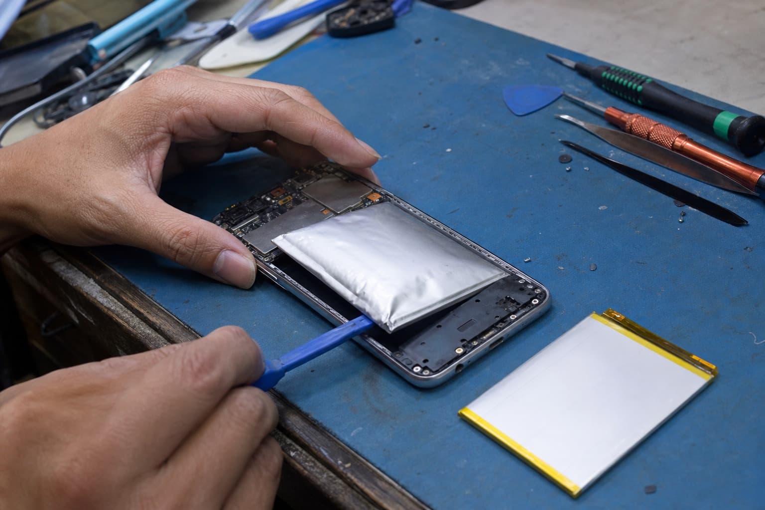 An Oppo-style slim Android phone opened on a repair bench with a slightly swollen battery being eased out by a plastic spudger, a flat replacement battery beside it on a blue anti-static mat, no logos