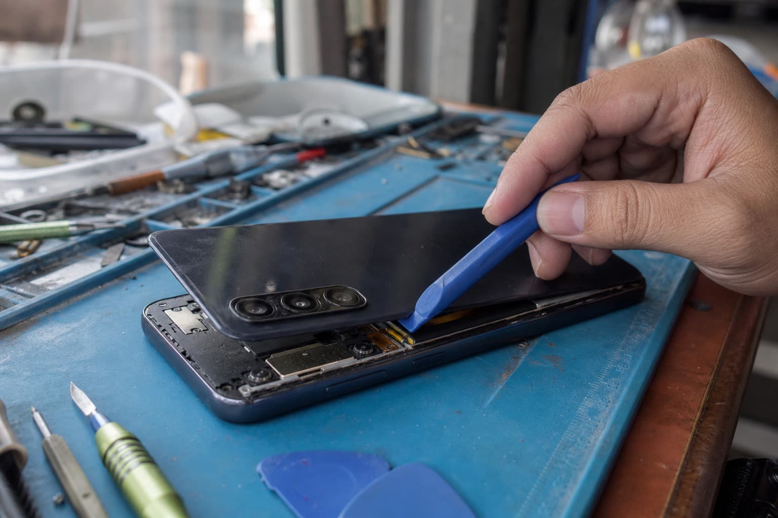 维修工作台上一部中端三星 Galaxy A 系列手机,掀开后盖露出三摄模组和内构,技师在蓝色防静电垫上用塑料撬棒检查,无 logo