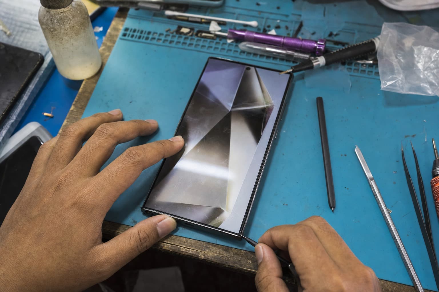 A premium Samsung Galaxy S Ultra-class flagship with a large flat AMOLED display and a slim stylus, powered on at a Malaysian repair-shop bench while a technician inspects the screen on a blue anti-static mat, no logos
