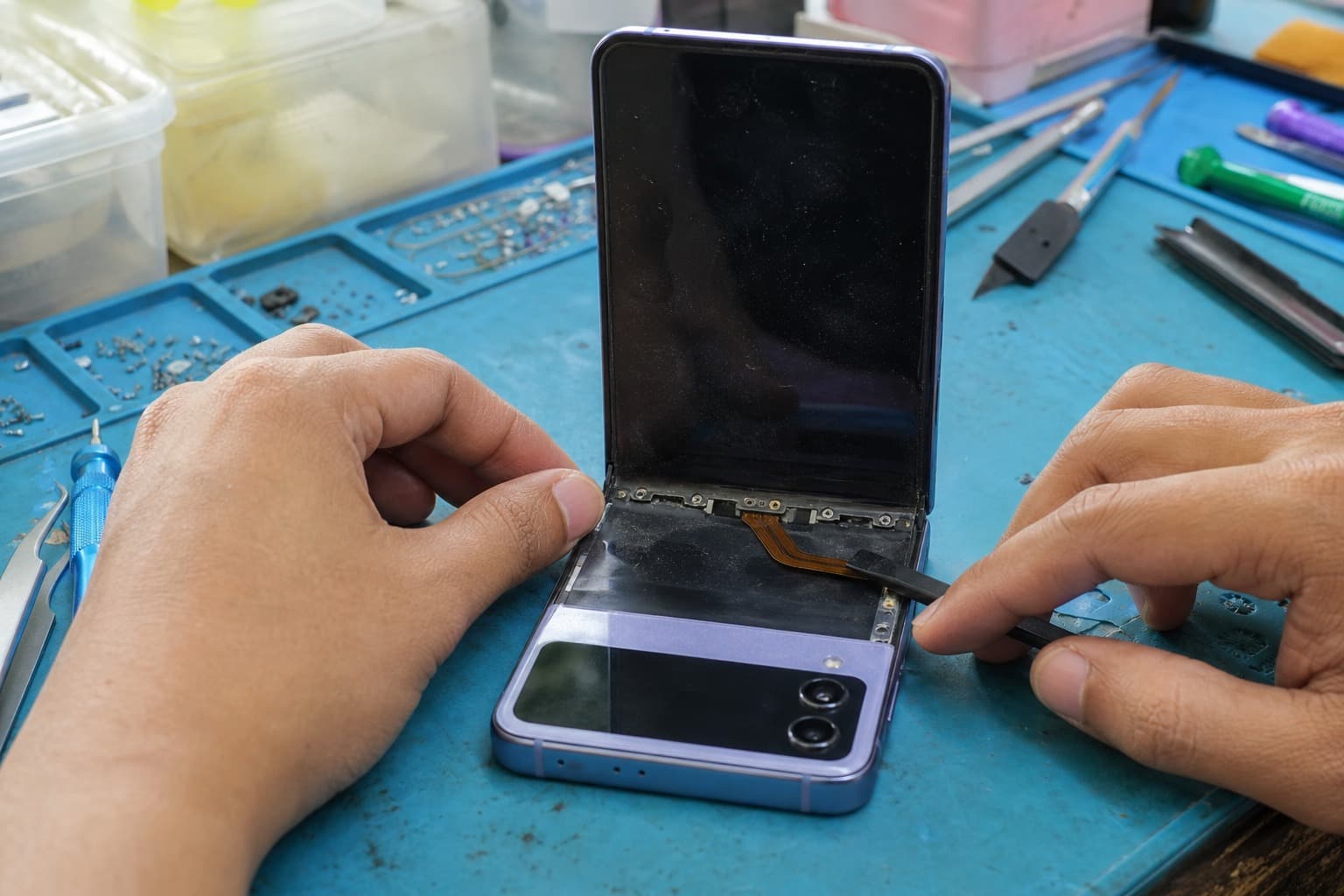 A Samsung Galaxy Z Flip-style clamshell foldable half-open on a repair bench, showing its tall inner folding screen and a small outer cover display, a technician inspecting the fold and flex cable with a spudger on a blue anti-static mat, no logos