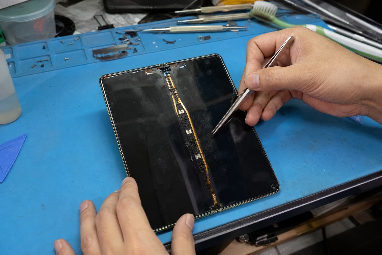 一部三星 Galaxy Z Fold 式折叠机摊平展开,露出大尺寸可弯曲内屏 AMOLED,技师在蓝色防静电垫上用精细镊子检查内屏折痕和穿过折叠边的柔性排线,无 logo
