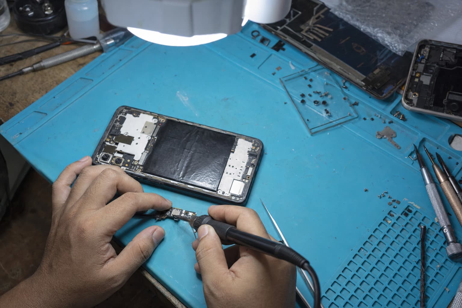 Telefon Android gaya Vivo dibuka di meja baiki dengan port pengecasan USB-C dibaiki menggunakan besi pateri mata halus di bawah lampu pembesar di atas pad anti-statik biru, tiada logo