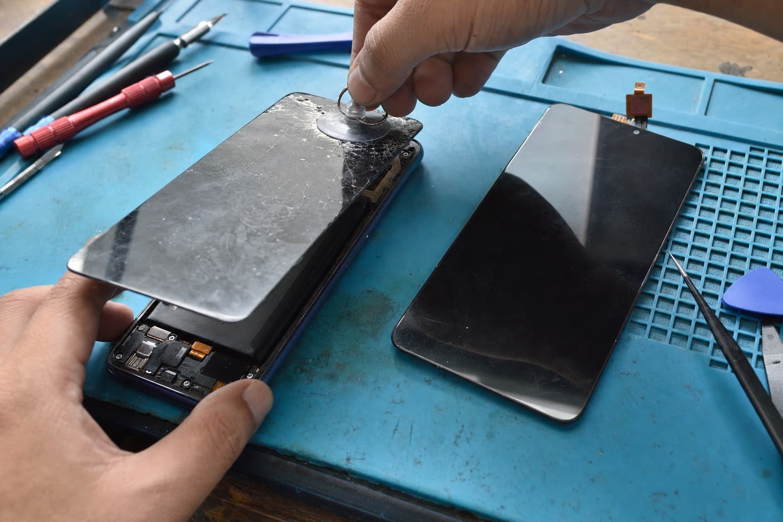 A Xiaomi/Redmi-style Android phone on a repair bench with its cracked front screen lifted off by a suction cup, a fresh replacement display lying beside it on a blue anti-static mat, no logos